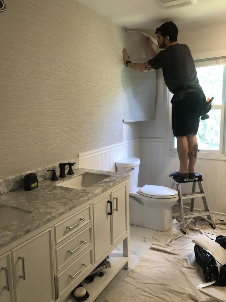 Professional wallpaper installer on a ladder carefully positioning a water resistant faux grasscloth texture wallpaper panel above wainscoting in a bathroom, Costa Blanca Spain